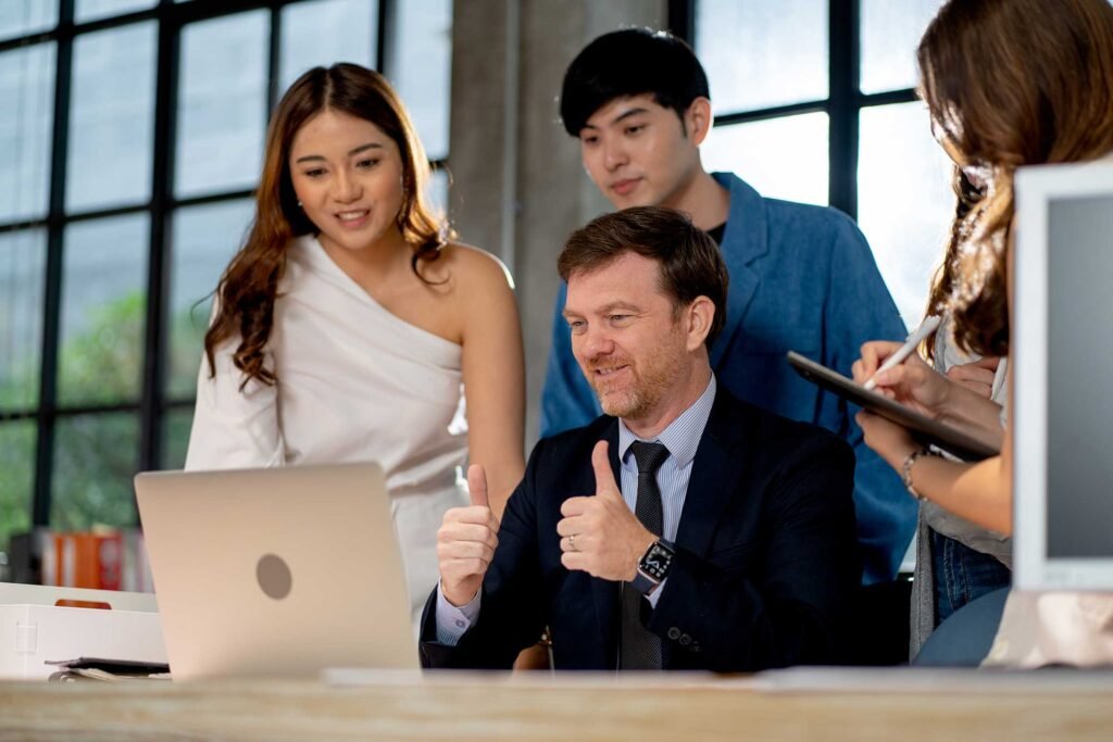 A group of professionals, including a man in a suit giving thumbs up, working together around a laptop in a modern office setting.